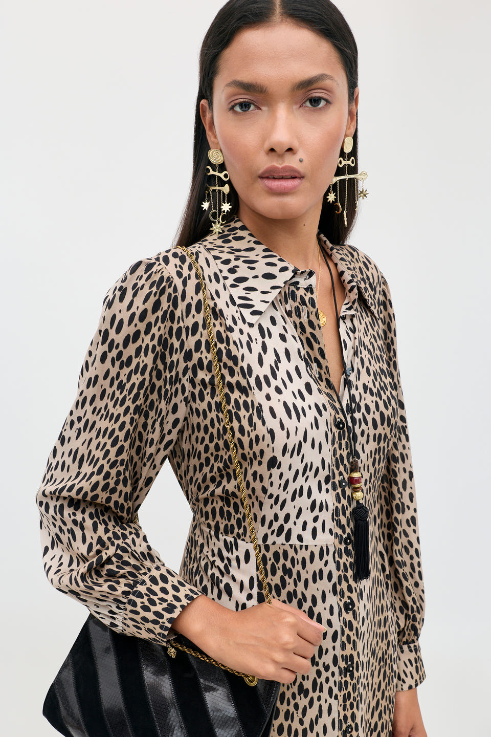 A petite woman stands against a plain background, effortlessly showcasing everyday style in the Maddison - Antelope Spot Black shirt dress, paired with large gold earrings and a black purse featuring a gold chain strap.