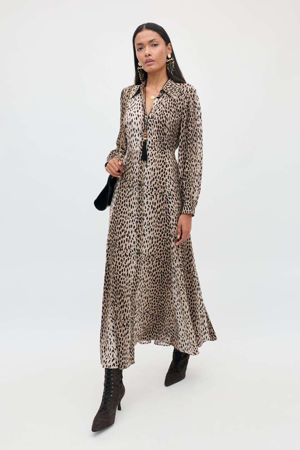 A petite woman stands against a plain white background, holding a black bag at her side and wearing the Maddison - Antelope Spot Black long-sleeve, ankle-length shirt dress with black boots and earrings—an effortless example of everyday style.