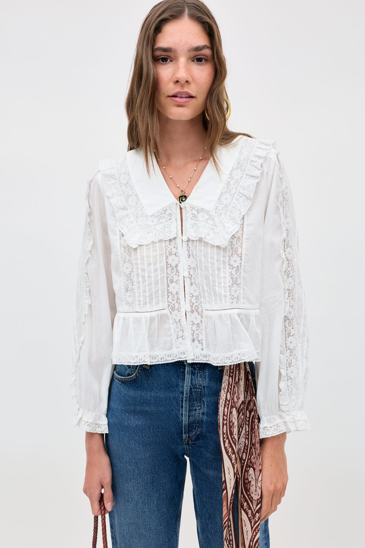 A woman wears the Maribelle - Ivory, a vintage-inspired white blouse with lace accents and a ruffled collar, paired with blue jeans. She stands against a plain background, holding a bag adorned with a patterned scarf.