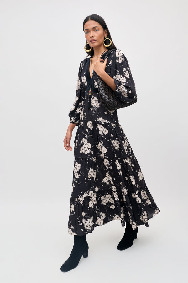 A woman wearing the Marlene - Daisy Field Black long-sleeve maxi dress with a deep V-neckline, paired with black boots, large gold hoop earrings, and a textured black shoulder bag, stands against a plain light background.