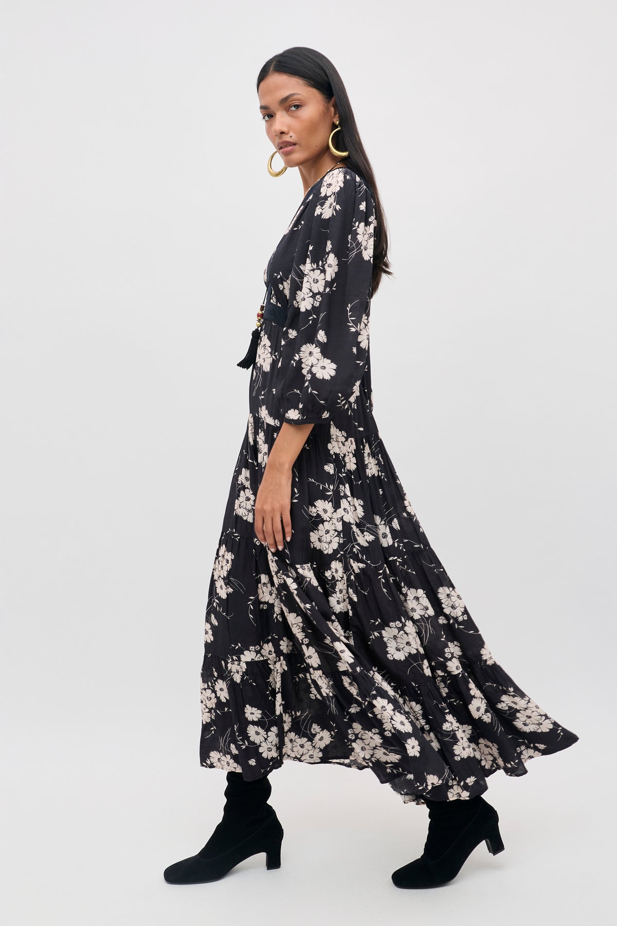 Woman wearing the Marlene - Daisy Field Black long, flowing dress with white floral pattern, styled with black boots and large hoop earrings, standing sideways against a plain light background.