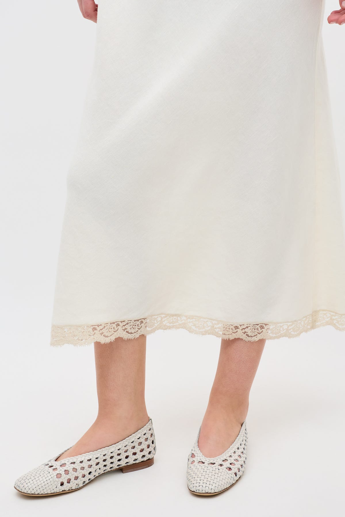 Someone is wearing the Marlia - Cream, a long off-white skirt with lace-trimmed hem, paired with white woven slip-on shoes and posing against a plain white background.