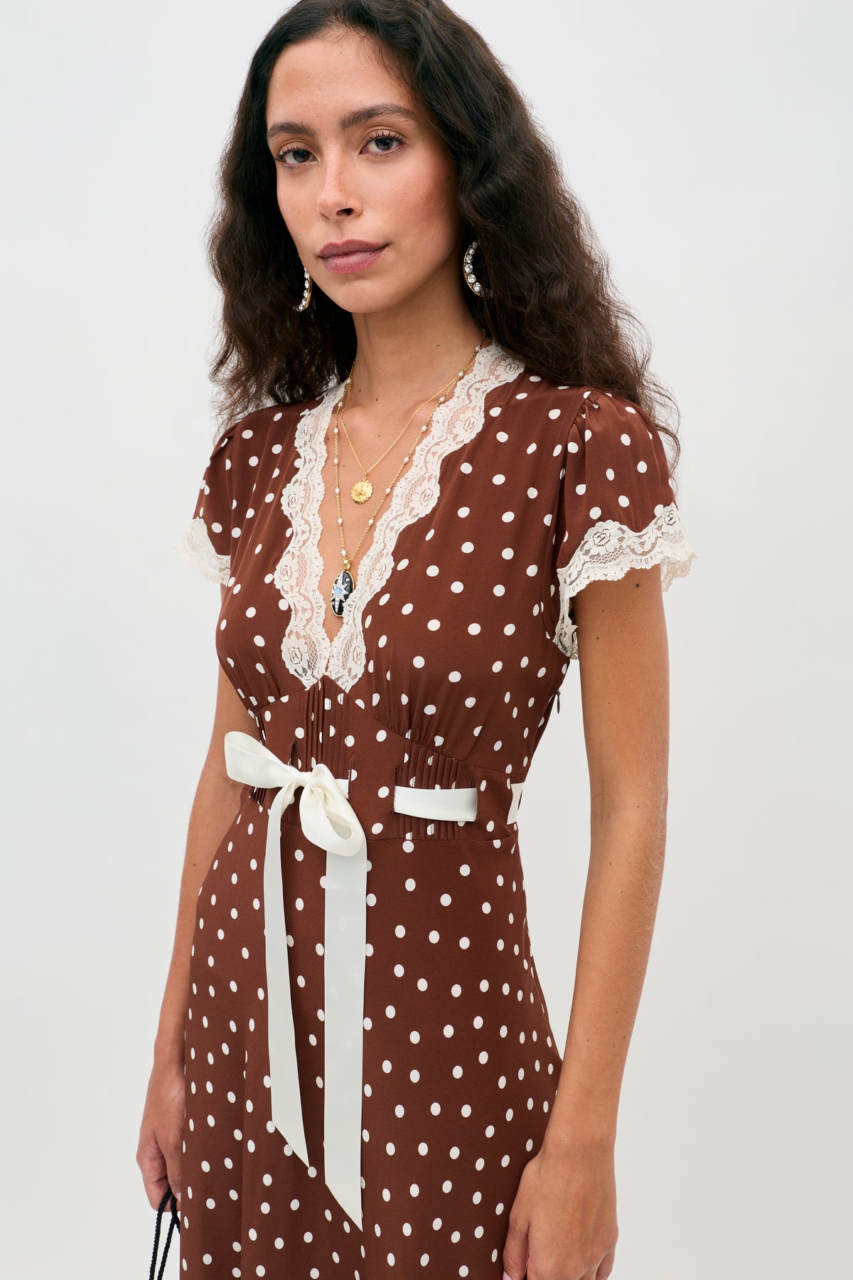 Wearing the Marlice - Layla Dot Brown dress, a woman with long curly hair stands against a plain light background. The dress features brown polka dots, lace trim, and a white ribbon belt. She completes her look with layered necklaces and hoop earrings.