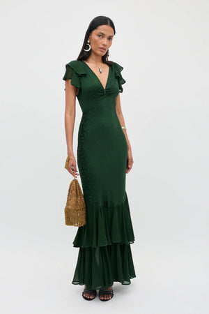 A woman stands against a plain light background, wearing the Marlow - Floral Trail Jacquard Dark Green gown—a floor-length dress with ruffled details and a deep V-neckline. She accessorizes with large hoop earrings and holds a woven gold handbag.