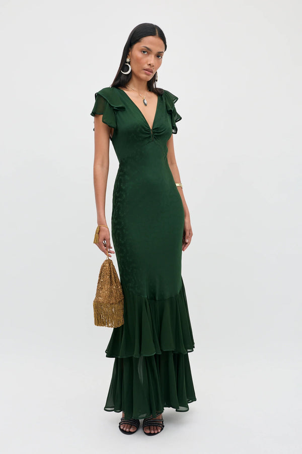 A woman stands against a plain light background, wearing the Marlow - Floral Trail Jacquard Dark Green gown—a floor-length dress with ruffled details and a deep V-neckline. She accessorizes with large hoop earrings and holds a woven gold handbag.
