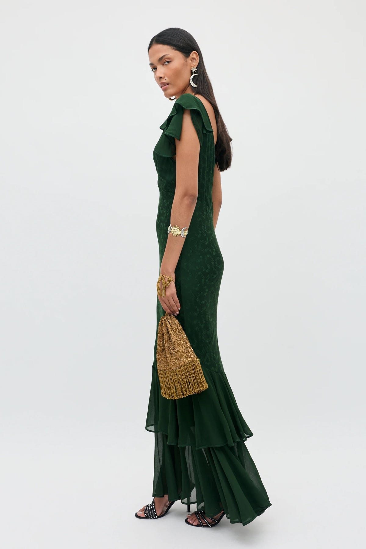 Dressed in the Marlow - Floral Trail Jacquard Dark Green, a woman holds a gold tasseled bag. She accessorizes with silver earrings, a bracelet, and black strappy shoes, standing against a plain light background.