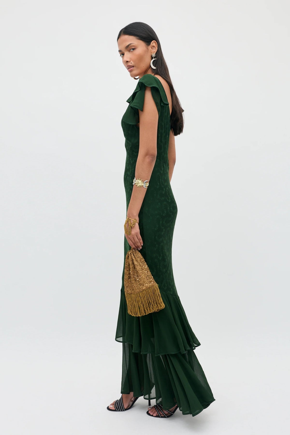 Dressed in the Marlow - Floral Trail Jacquard Dark Green, a woman holds a gold tasseled bag. She accessorizes with silver earrings, a bracelet, and black strappy shoes, standing against a plain light background.