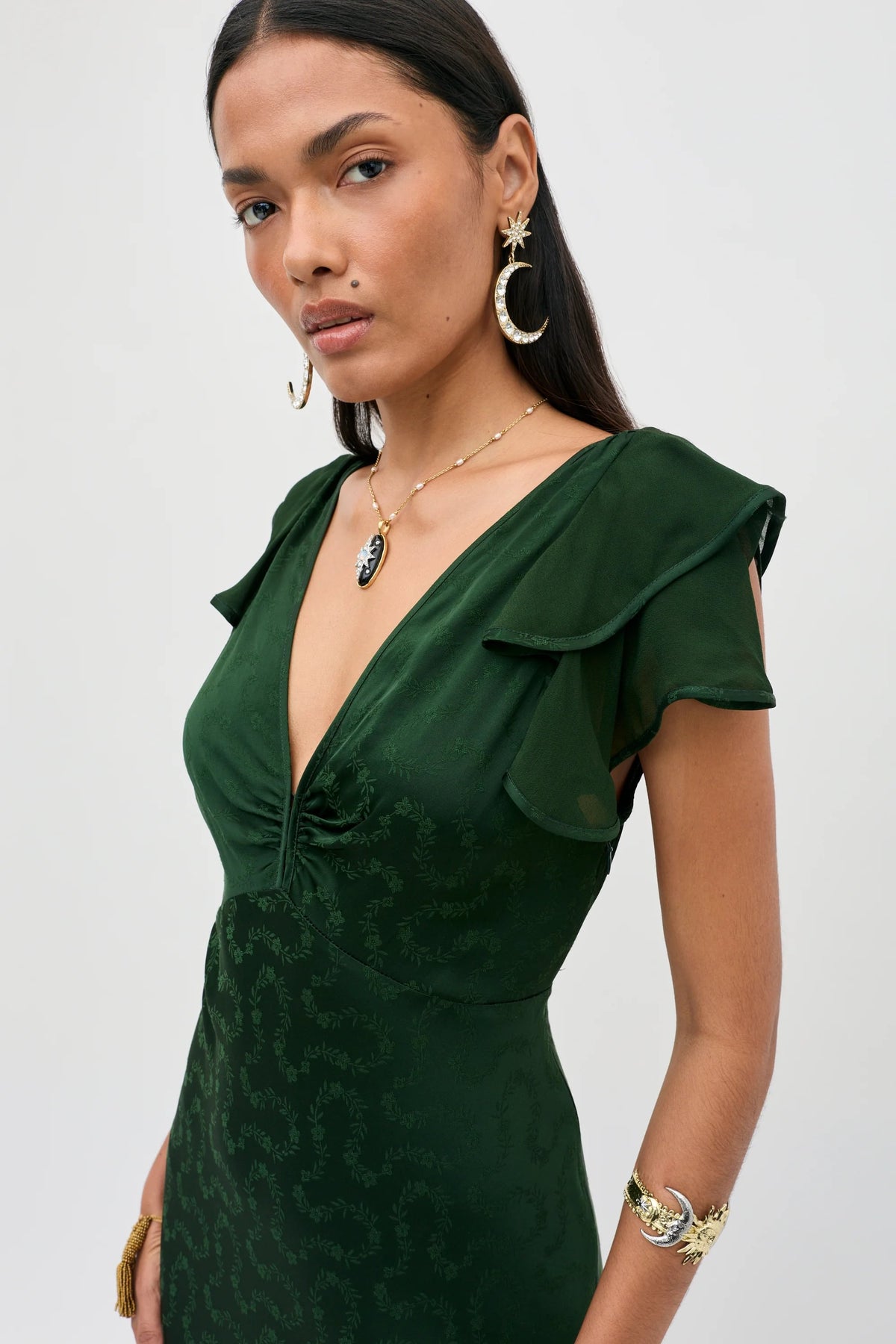 A woman stands against a plain light background wearing the Marlow - Floral Trail Jacquard Dark Green dress, which features short sleeves and ruffled shoulders. She accessorizes with gold jewelry, including large moon-shaped earrings and a pendant necklace.