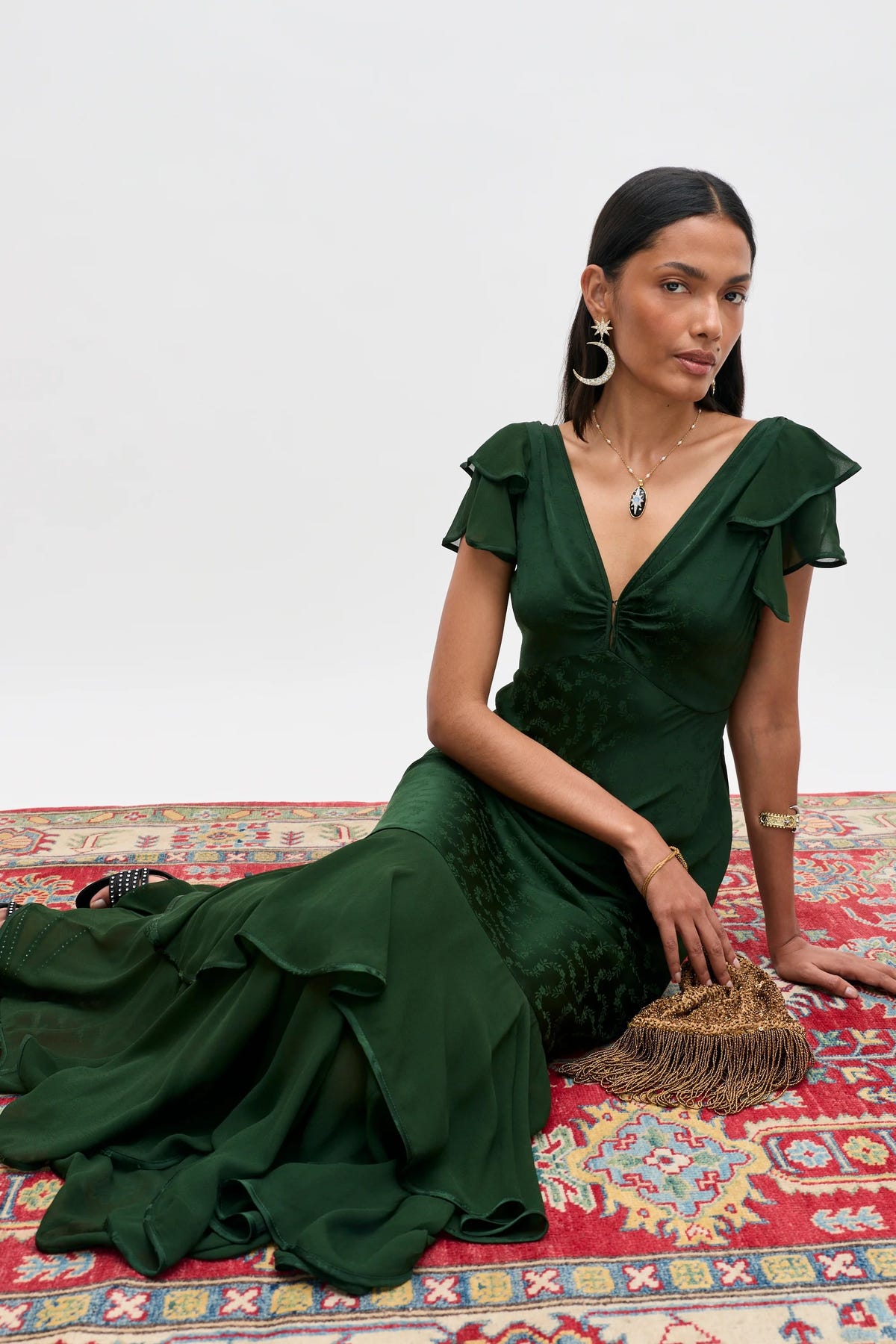 A woman sits on a colorful patterned rug, wearing the Marlow – Floral Trail Jacquard Dark Green dress, a deep green, ruffled, floor-length gown. She accessorizes with statement earrings, a necklace, gold bracelets, and has a gold fringed clutch by her side. The background is white.