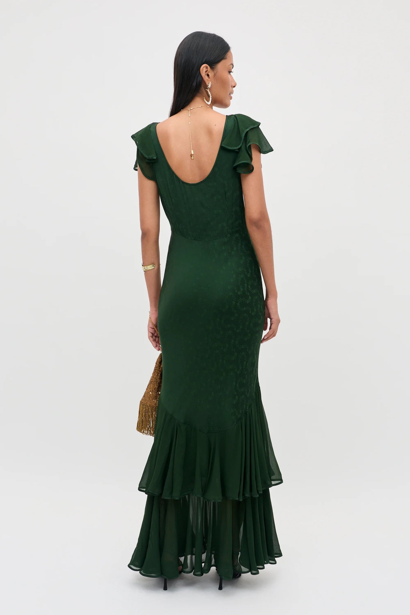 A woman stands facing away in the Marlow - Floral Trail Jacquard Dark Green floor-length dress, featuring ruffled detailing. She holds a gold bag and accessorizes with long dark hair, gold earrings, and a gold bracelet against a plain light background.