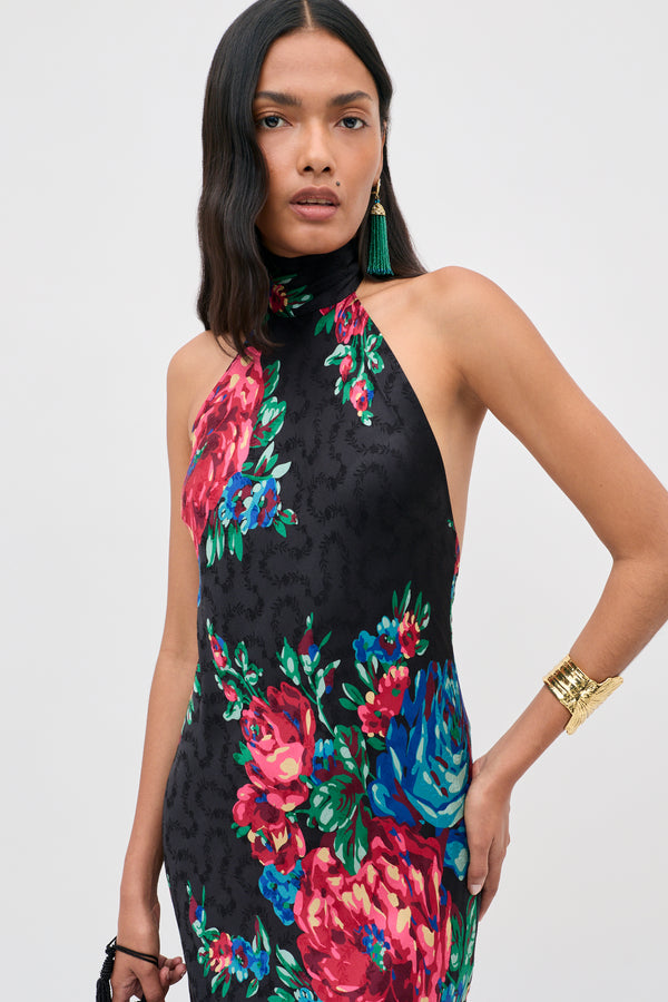 A woman models the Maura - Vintage Bouquet Black dress, a sleeveless black piece featuring large, colorful floral patterns. She accessorizes with a gold bracelet, green tassel earrings, and carries a small black bag. Her long, dark wavy hair frames her face as she gazes confidently at the camera.