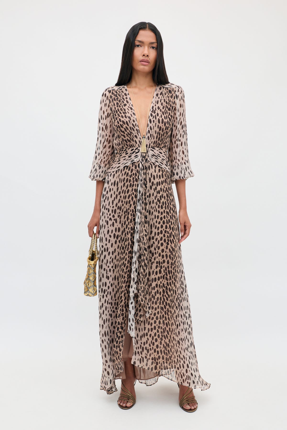 A woman stands against a white background wearing the Meera - Antelope Spot Black, a long, flowing crinkled-chiffon gown with a deep V-neck animal print design, holding a small gold purse and wearing strappy sandals.