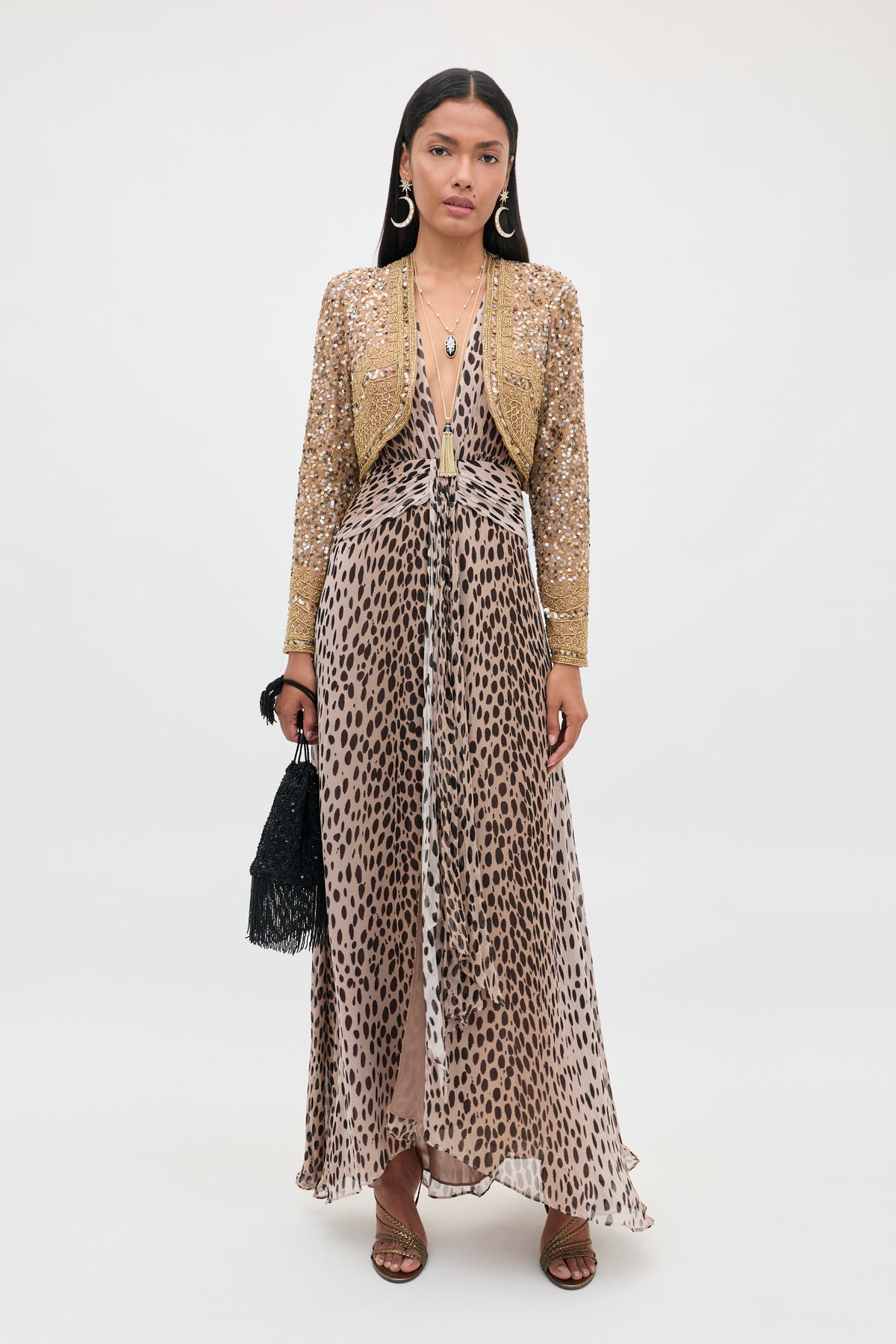 A woman stands against a plain white background, wearing the “Meera - Antelope Spot Black” leopard-print maxi dress with a plunging neckline, layered beneath a gold sequin jacket. She accessorizes with hoop earrings, long dark hair styled loose, strappy sandals, and holds a black fringe bag.