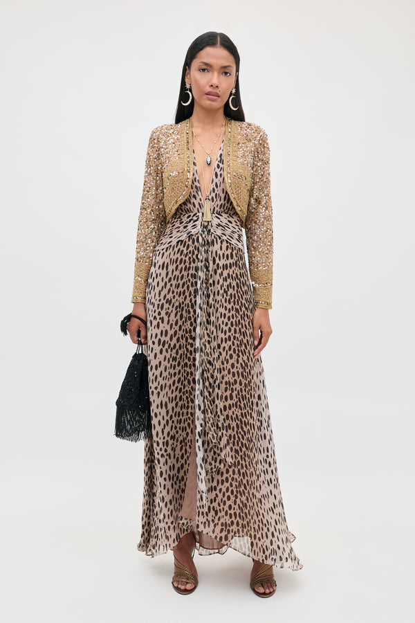 A woman stands against a plain white background, wearing the “Meera - Antelope Spot Black” leopard-print maxi dress with a plunging neckline, layered beneath a gold sequin jacket. She accessorizes with hoop earrings, long dark hair styled loose, strappy sandals, and holds a black fringe bag.