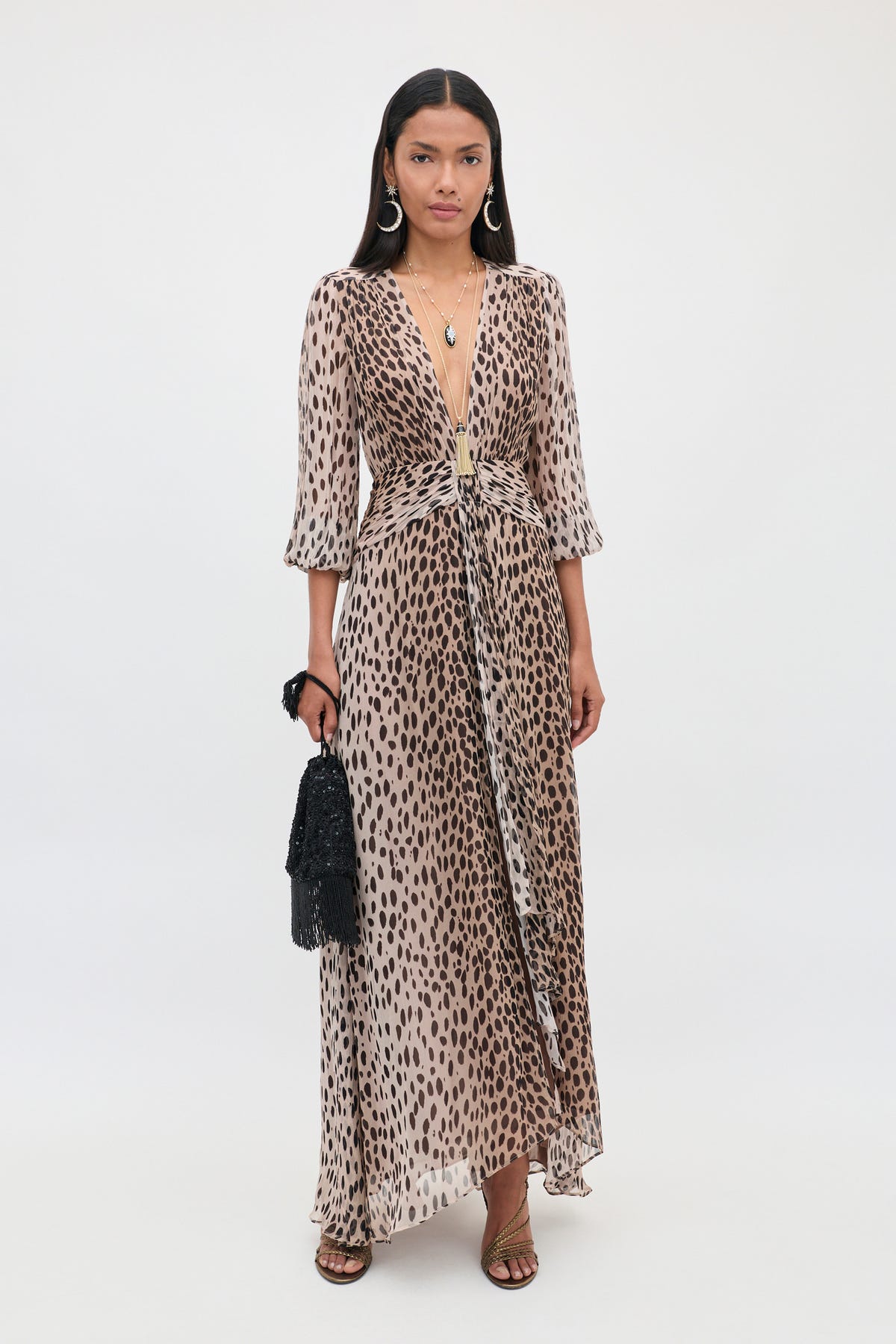 A woman with long dark hair stands against a plain white background wearing the Meera - Antelope Spot Black dress—an elegant, long dress featuring a deep V neckline and antelope spot print. She accessorizes with sandals, hoop earrings, layered necklaces, and holds a black handbag.