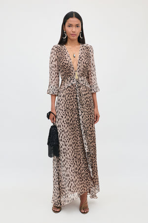 A woman with long dark hair stands against a plain white background wearing the Meera - Antelope Spot Black dress—an elegant, long dress featuring a deep V neckline and antelope spot print. She accessorizes with sandals, hoop earrings, layered necklaces, and holds a black handbag.