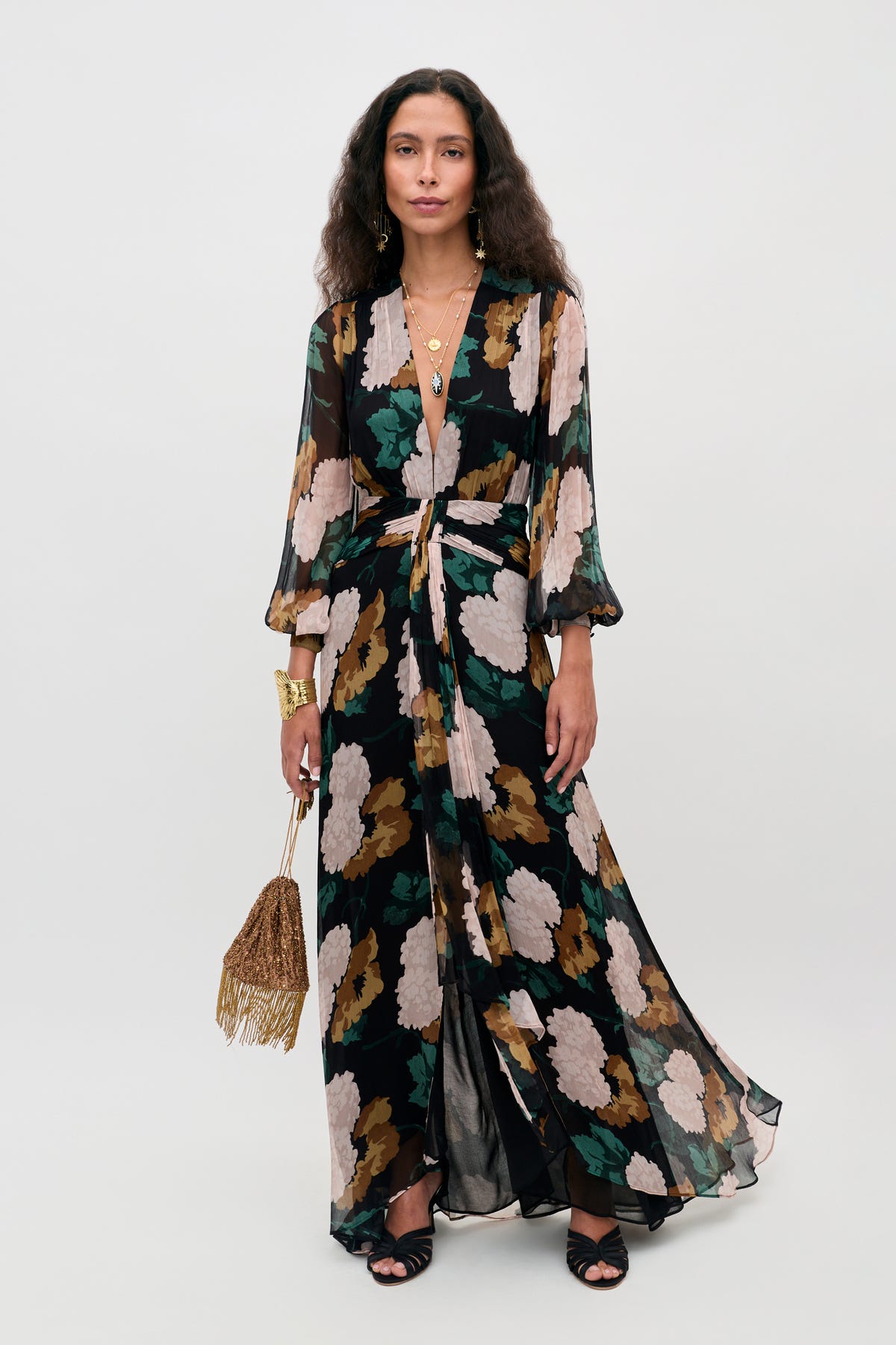 A woman wearing the Meera - Hydrangea Floral Charcoal dress—a long, flowy dress featuring black, tan, green, and cream florals with sheer sleeves—carries a fringed gold handbag and accessorizes with layered necklaces and bracelets.