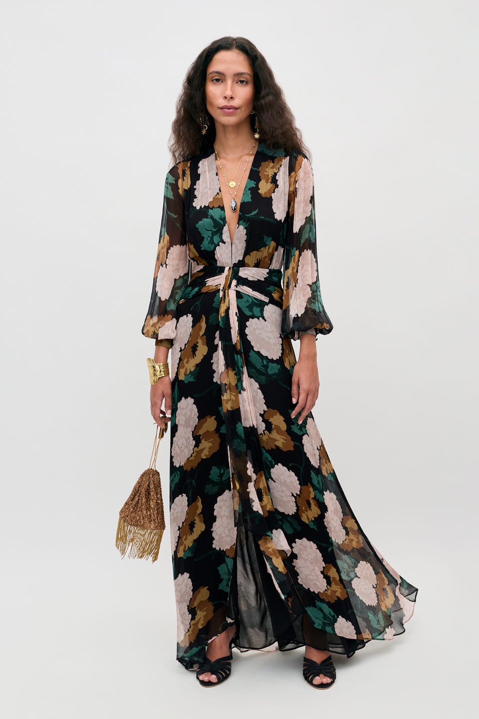 A woman wearing the Meera - Hydrangea Floral Charcoal dress—a long, flowy dress featuring black, tan, green, and cream florals with sheer sleeves—carries a fringed gold handbag and accessorizes with layered necklaces and bracelets.