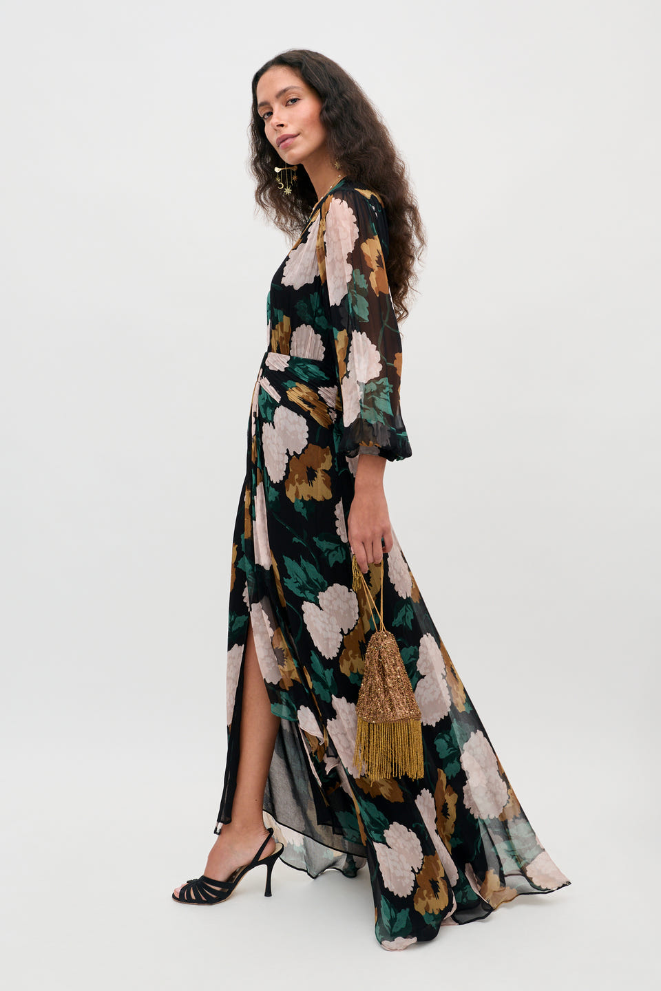 A woman wearing the Meera – Hydrangea Floral Charcoal dress with a long, flowy silhouette and slit, holding a gold tassel bag, styled with black heeled sandals, poses against a plain light background.