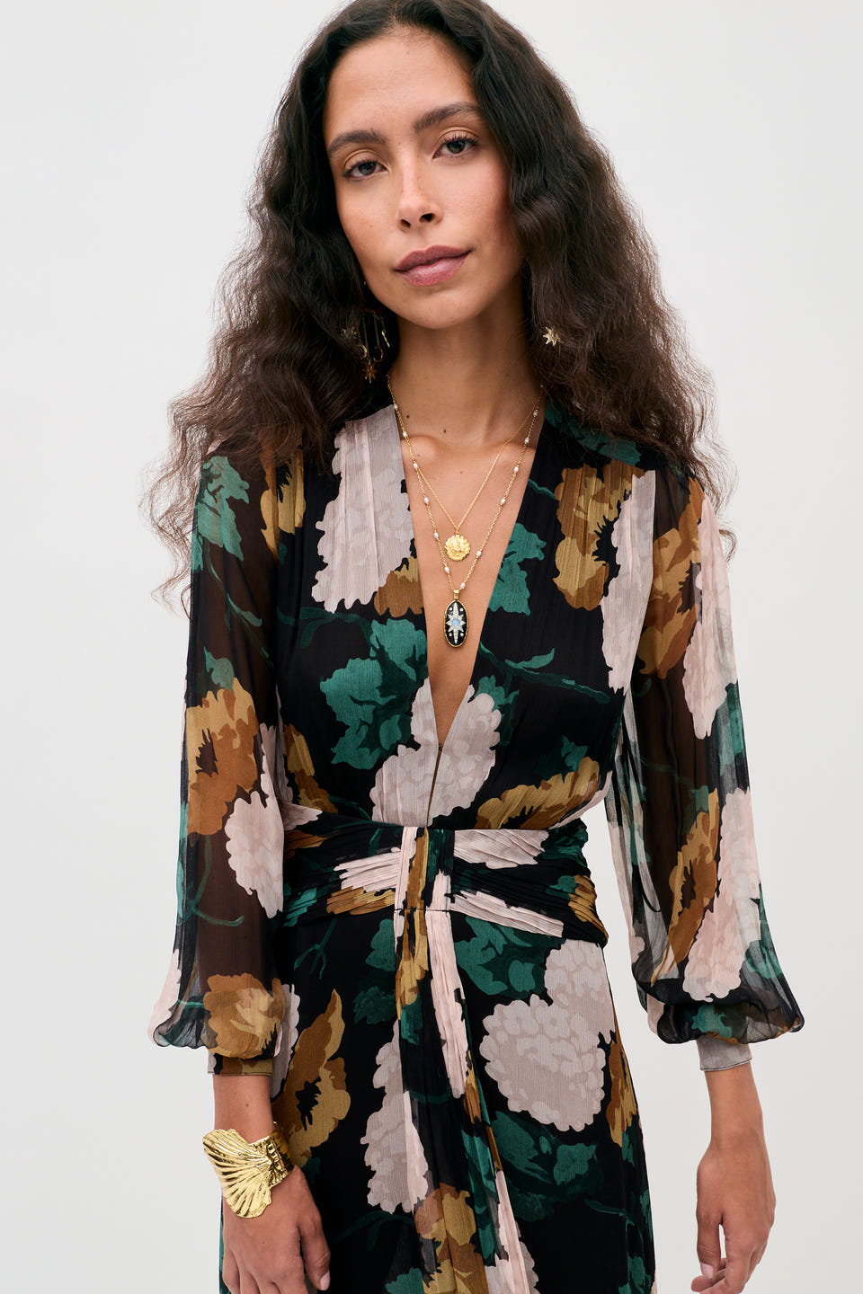A woman with long wavy hair wears the Meera - Hydrangea Floral Charcoal dress, featuring a floral pattern and a deep neckline. She accessorizes with layered gold necklaces, gold earrings, and a gold bracelet, standing against a plain light background.