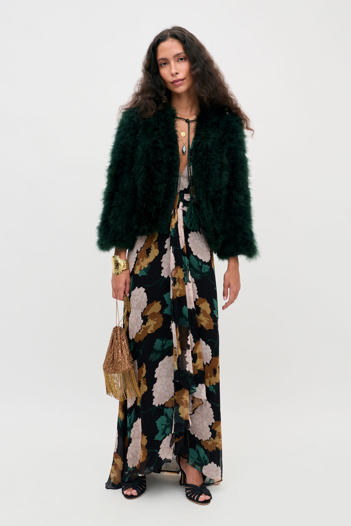 A woman wears a Vira - Dark Green feathered jacket over a floral maxi dress with a gold fringe bag.