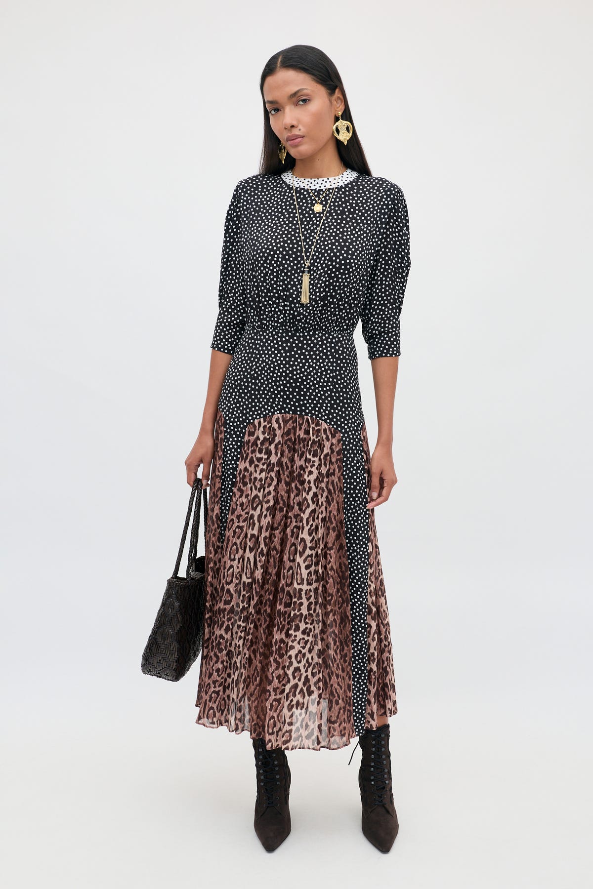 A woman stands against a plain background wearing the “meg - leopard polka dot” set: a black-and-white polka dot top paired with a long pleated midi skirt featuring bold leopard print panels. She completes the look with black boots, gold jewelry, and carries a black textured handbag.