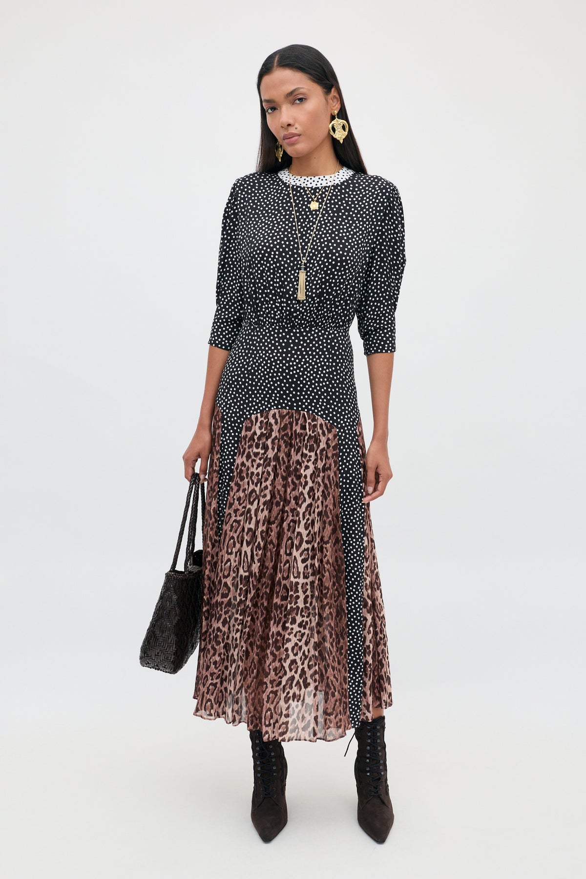 A woman stands against a plain background wearing the “meg - leopard polka dot” set: a black-and-white polka dot top paired with a long pleated midi skirt featuring bold leopard print panels. She completes the look with black boots, gold jewelry, and carries a black textured handbag.