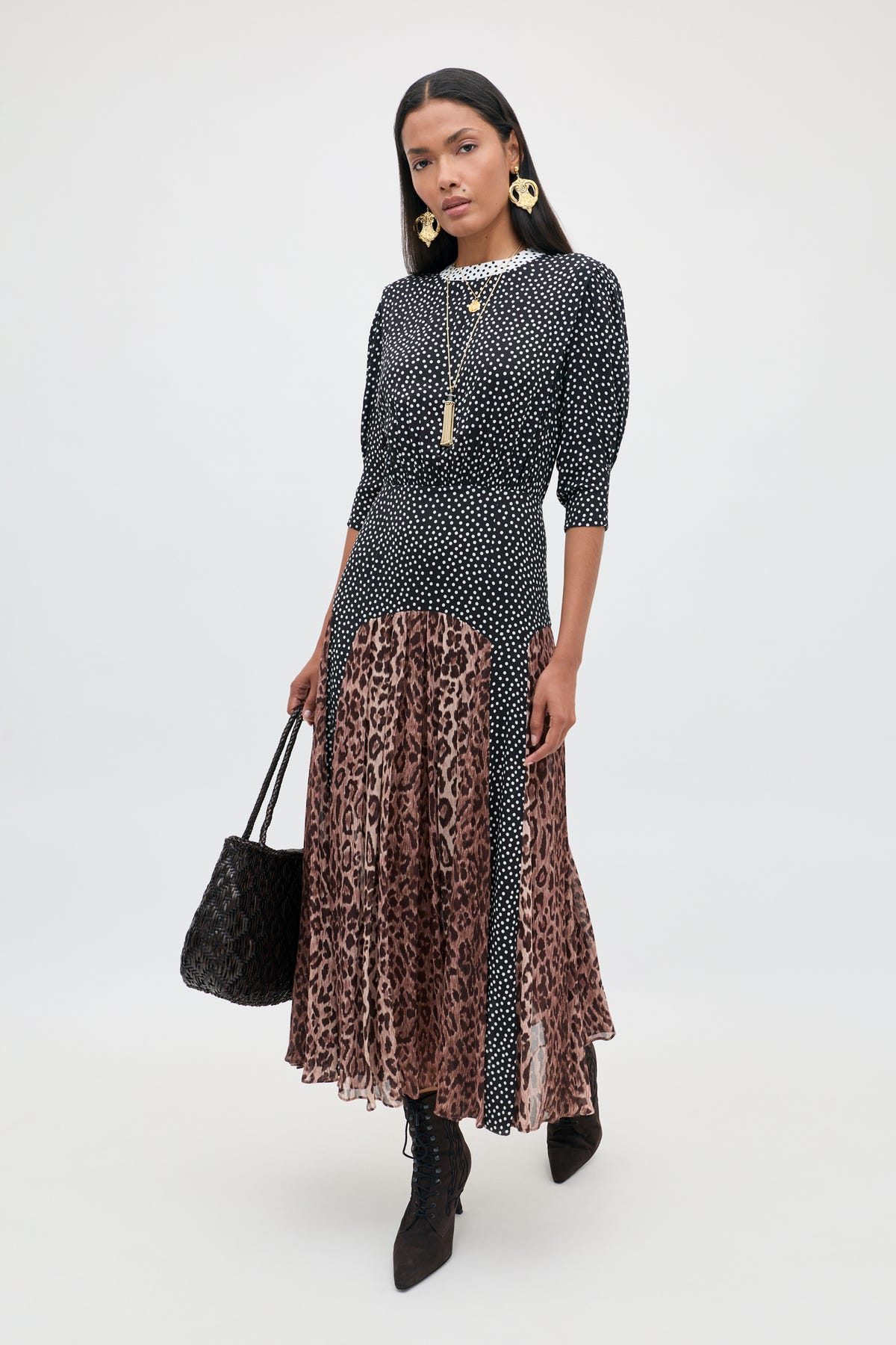 A woman stands against a plain light background, wearing the “meg - leopard polka dot” dress—a stylish crepe piece featuring a black and white polka-dot top and a brown leopard print midi skirt. She pairs the look with black boots, gold jewelry, and carries a black handbag.