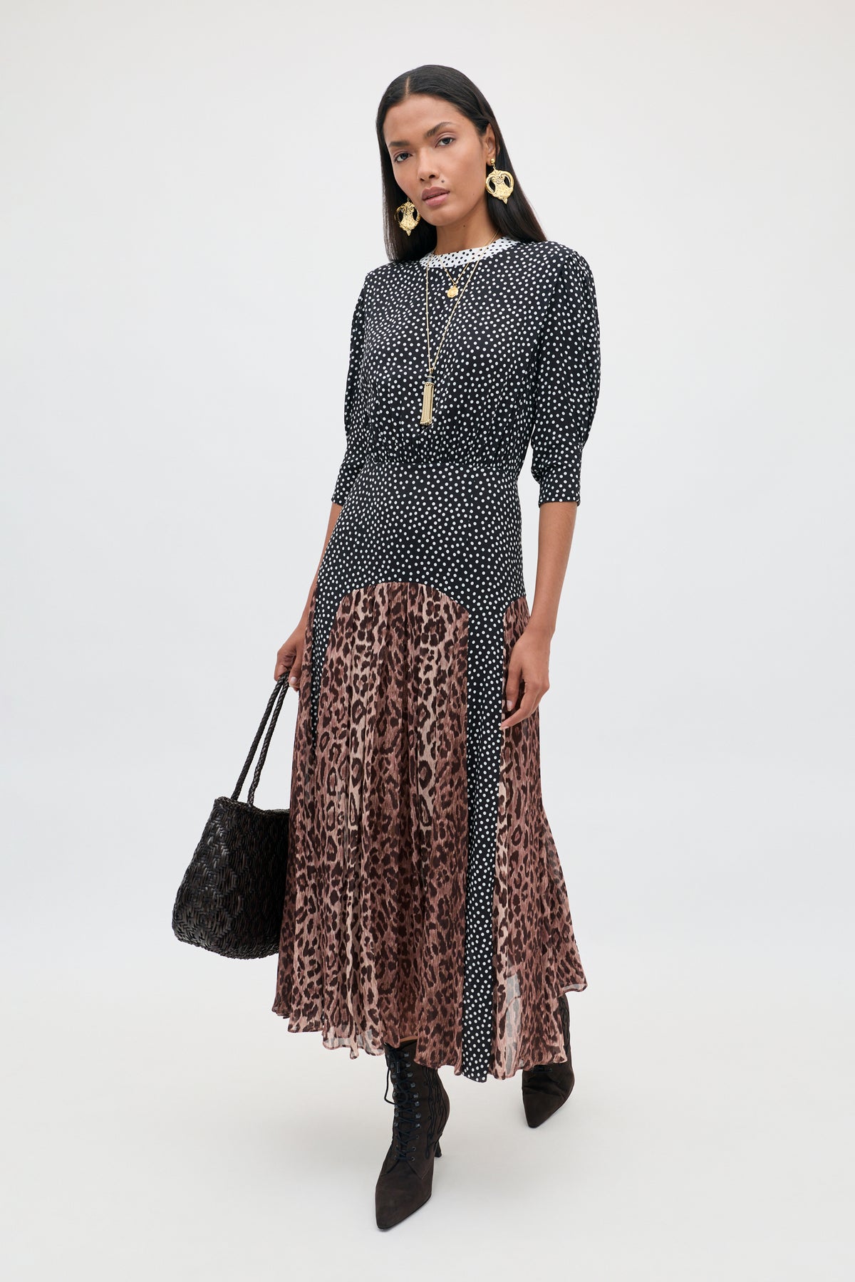 A woman stands against a plain light background, wearing the “meg - leopard polka dot” dress—a stylish crepe piece featuring a black and white polka-dot top and a brown leopard print midi skirt. She pairs the look with black boots, gold jewelry, and carries a black handbag.