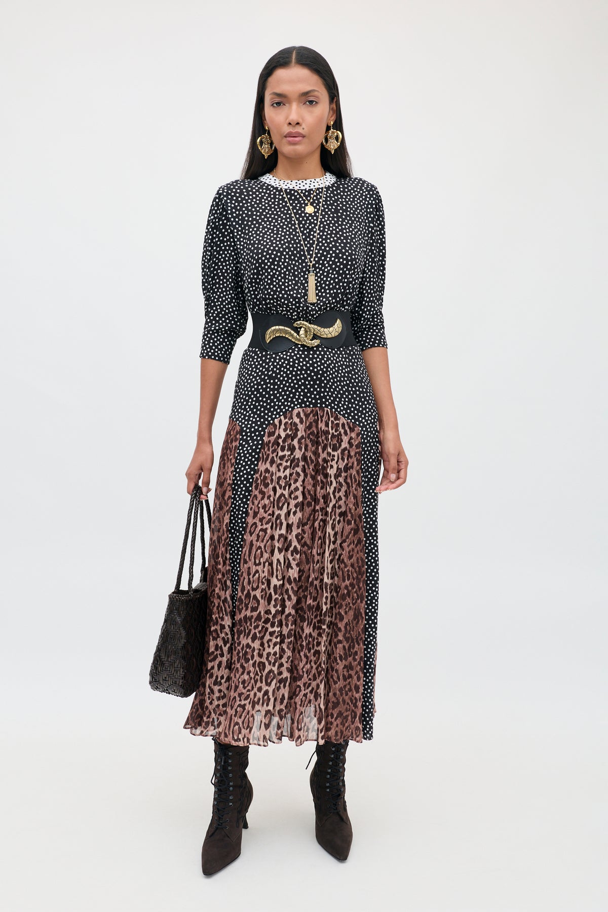 A woman stands against a plain background wearing the meg - leopard polka dot top paired with a pleated brown leopard print midi skirt, accessorized with a black belt, gold jewelry, black ankle boots, and holding a black textured bag.
