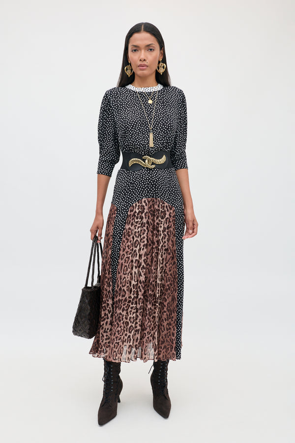 A woman stands against a plain background wearing the meg - leopard polka dot top paired with a pleated brown leopard print midi skirt, accessorized with a black belt, gold jewelry, black ankle boots, and holding a black textured bag.