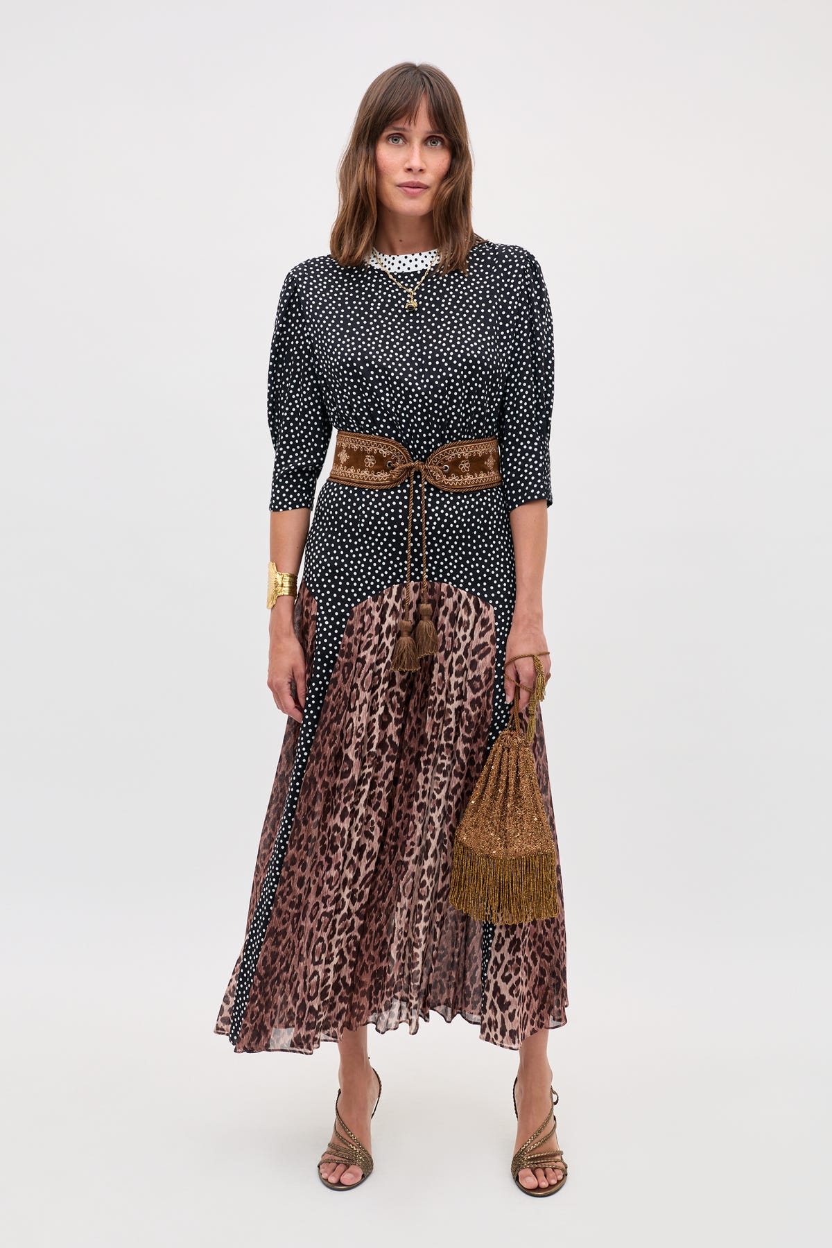 A woman models the "meg - leopard polka dot" look, pairing a black and white polka dot top with a brown leopard print pleated midi skirt, accessorized with gold sandals, chunky jewelry, and a brown fringed handbag.
