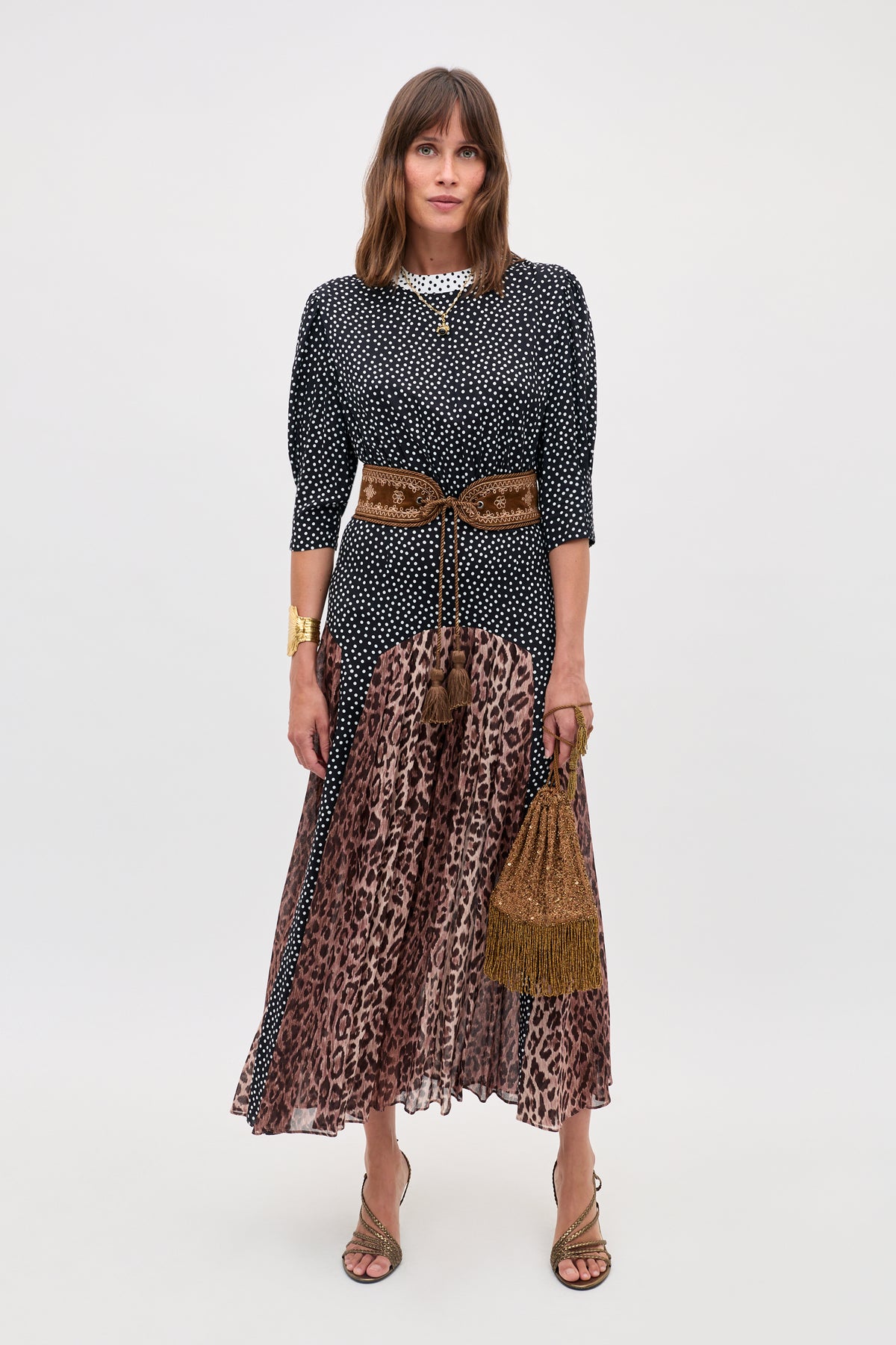 A woman models the "meg - leopard polka dot" look, pairing a black and white polka dot top with a brown leopard print pleated midi skirt, accessorized with gold sandals, chunky jewelry, and a brown fringed handbag.