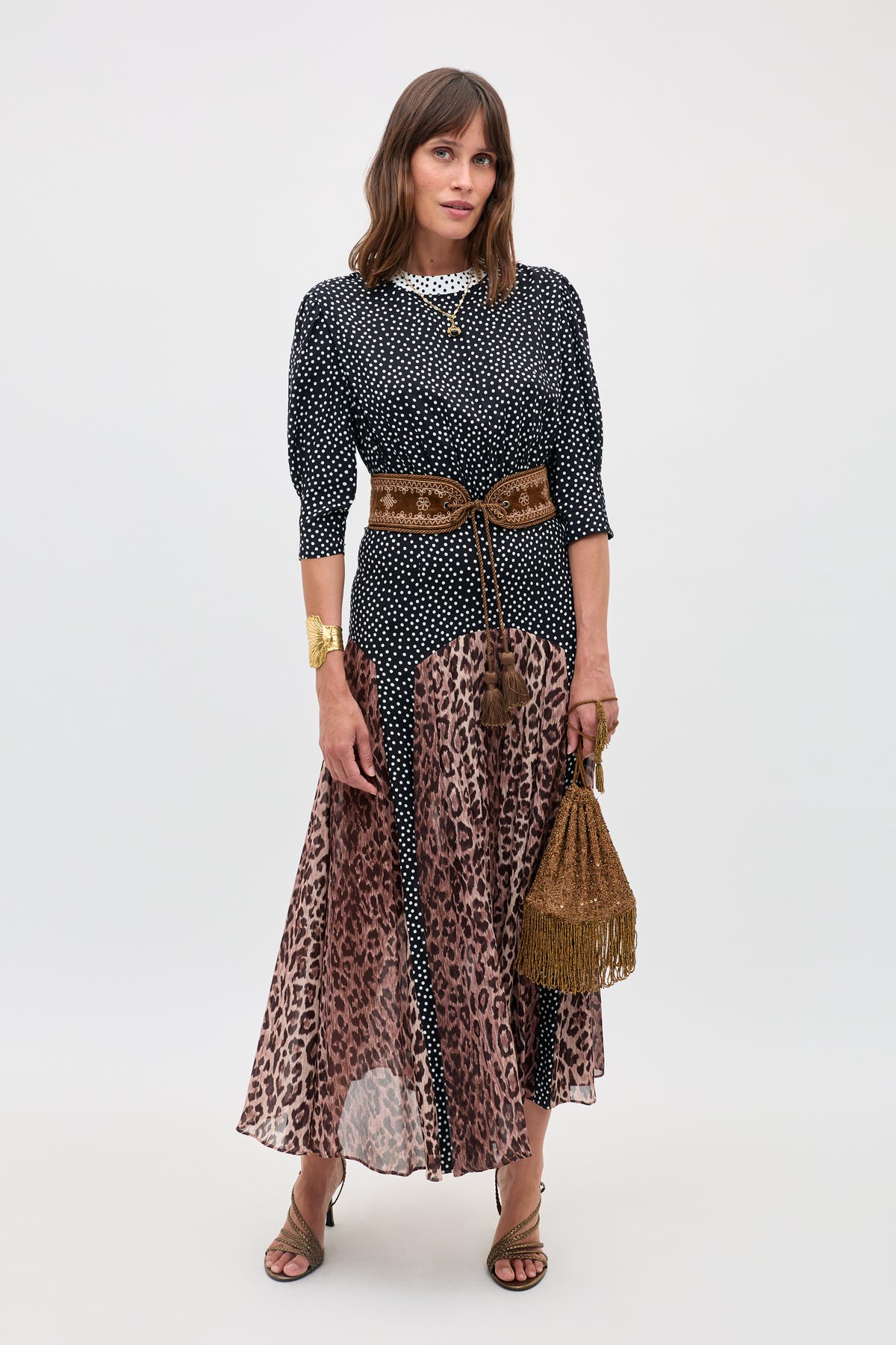 A woman stands against a plain light background wearing the "meg - leopard polka dot" top and skirt, paired with heeled sandals, a wide brown belt, and holding a brown fringed bag.