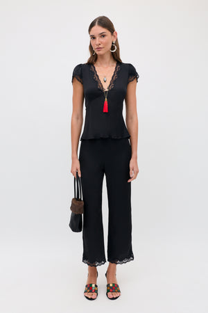 Woman standing against a plain background, wearing the Mireille - Black lace-trimmed top and pants, colorful sandals, hoop earrings, layered necklaces with a red tassel, and holding a black fur-trimmed bag.
