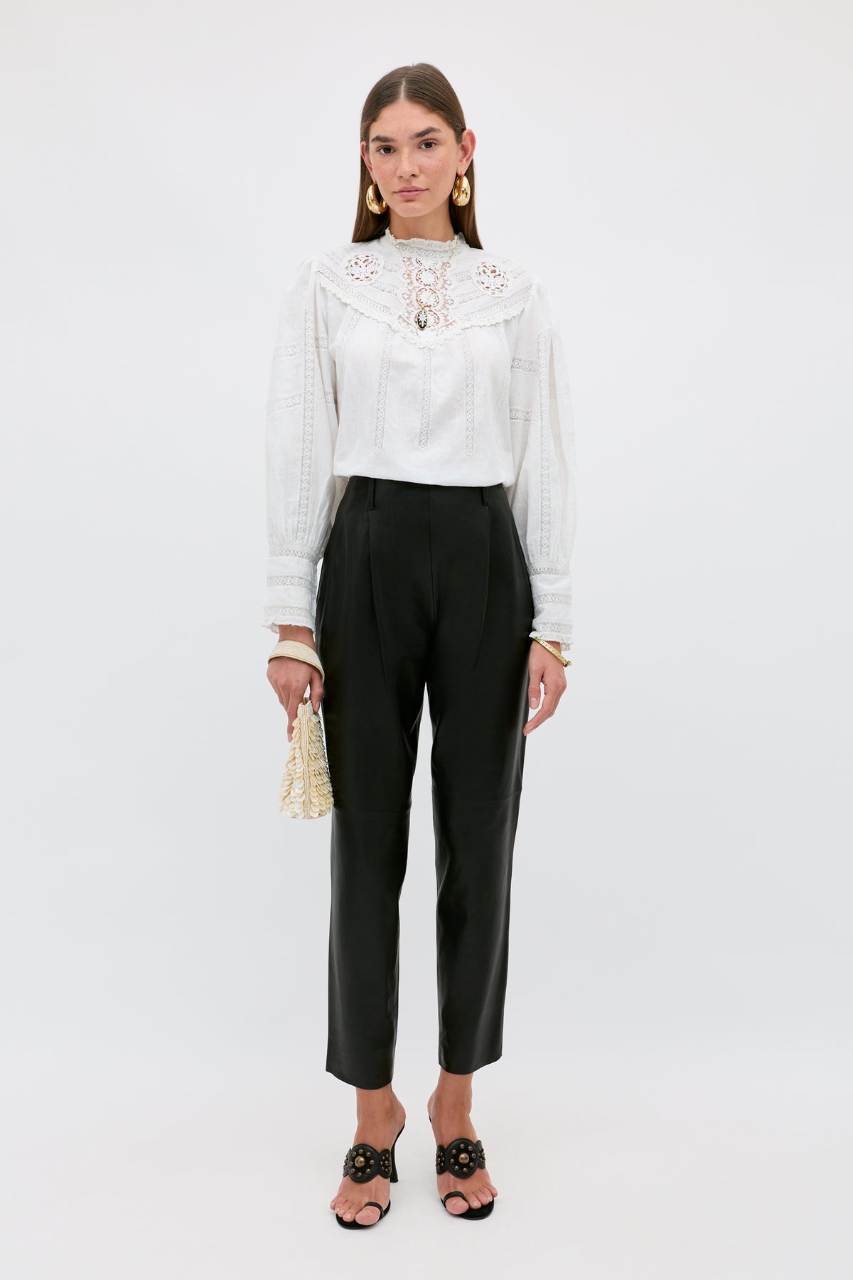 A woman stands against a plain background wearing the Nori - Charcoal ankle-length tailored pants, paired with a white long-sleeve lace blouse, black sandals, gold earrings, and holding a small raffia bag.