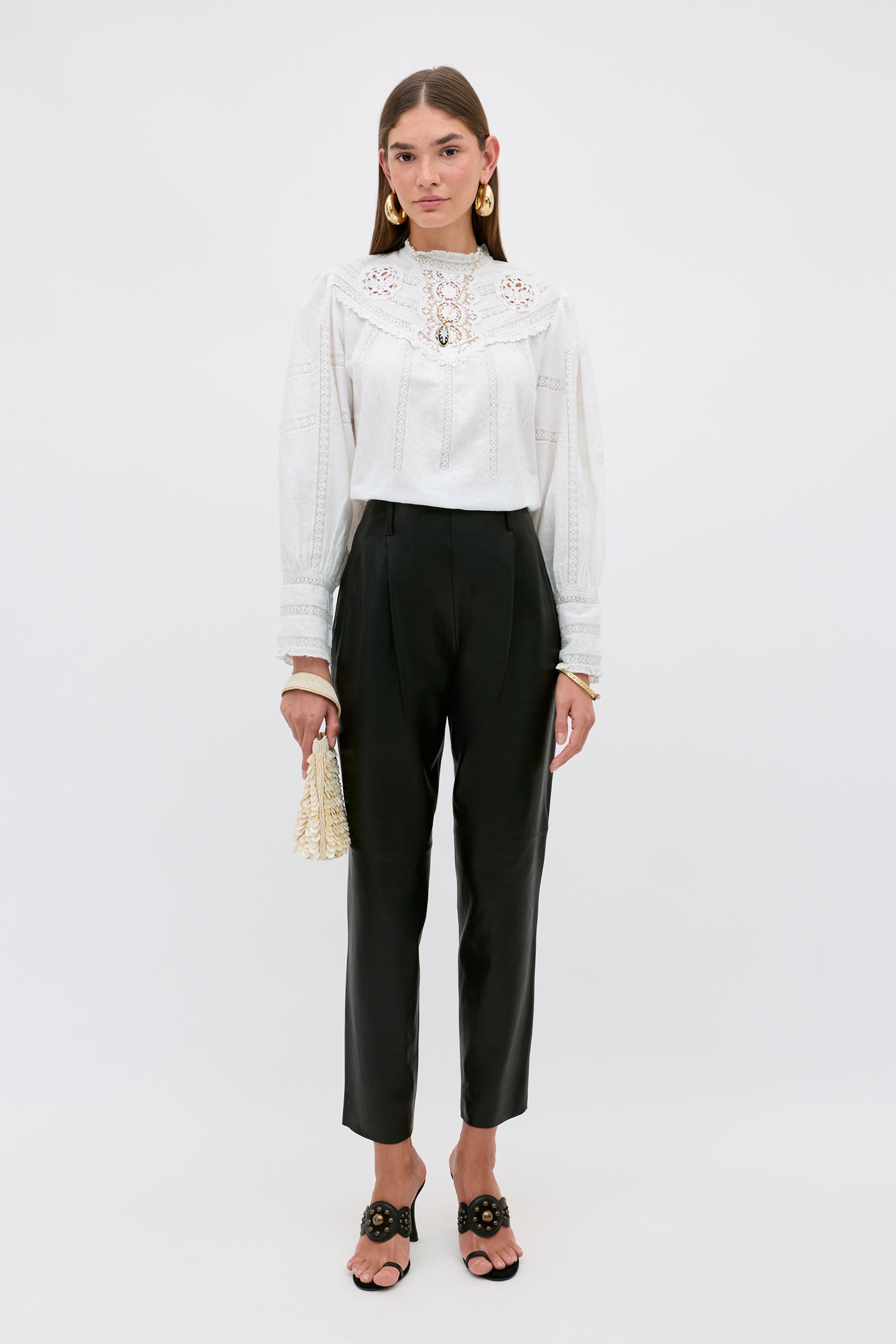 A woman stands against a plain background wearing the Nori - Charcoal ankle-length tailored pants, paired with a white long-sleeve lace blouse, black sandals, gold earrings, and holding a small raffia bag.