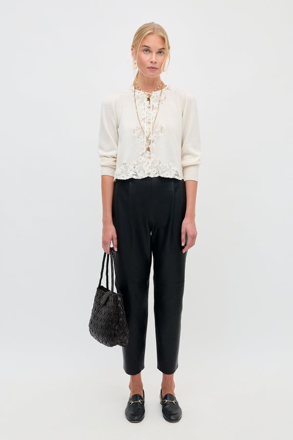A woman stands against a plain background wearing a cream lace top, black high-waisted pants, and black loafers, holding the Nori - Charcoal textured handbag. Her blonde hair is tied back and she has a neutral expression.