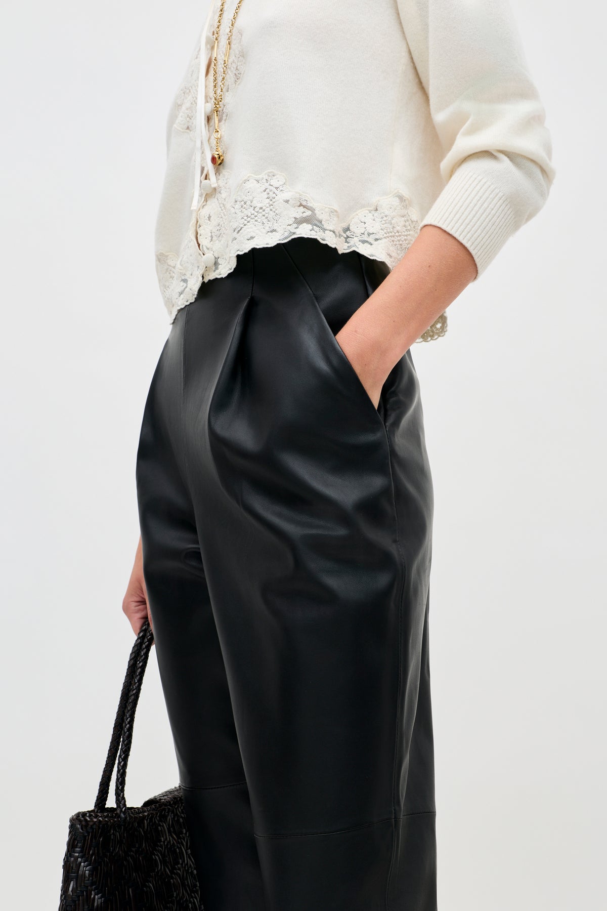 A person wearing the Nori - Charcoal sweater with white lace trim and high-waisted black leather pants, hands in pockets and holding a black woven handbag. The photo is cropped at the shoulders and knees.