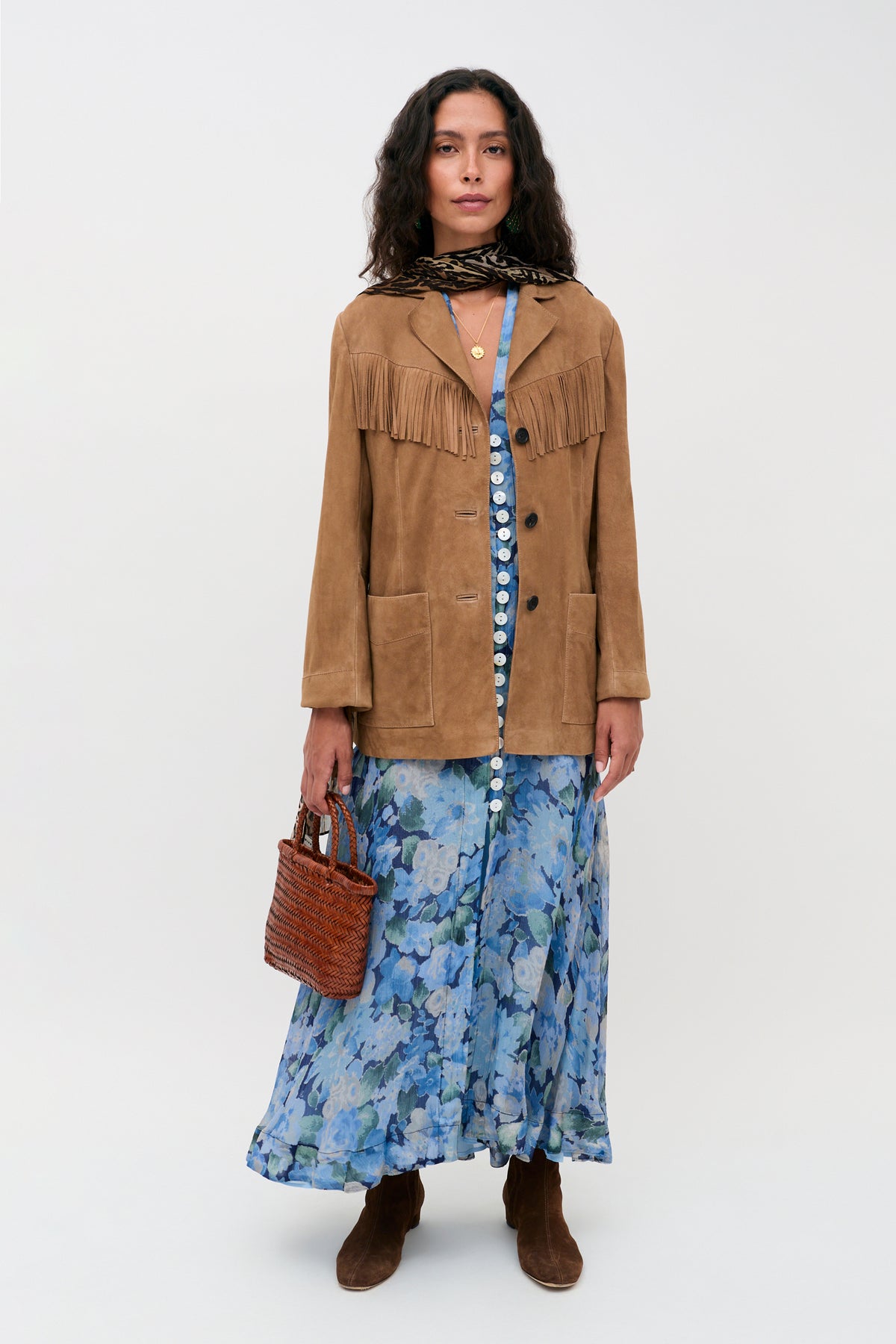 A woman wearing the Novara Dress in Chelsea Floral Blue, styled with a brown fringe jacket, brown boots, a scarf, and holding a woven bag stands against a plain background.