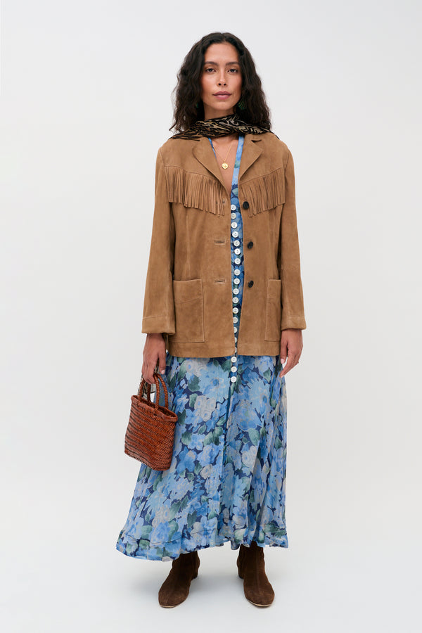 A woman wearing the Novara Dress in Chelsea Floral Blue, styled with a brown fringe jacket, brown boots, a scarf, and holding a woven bag stands against a plain background.