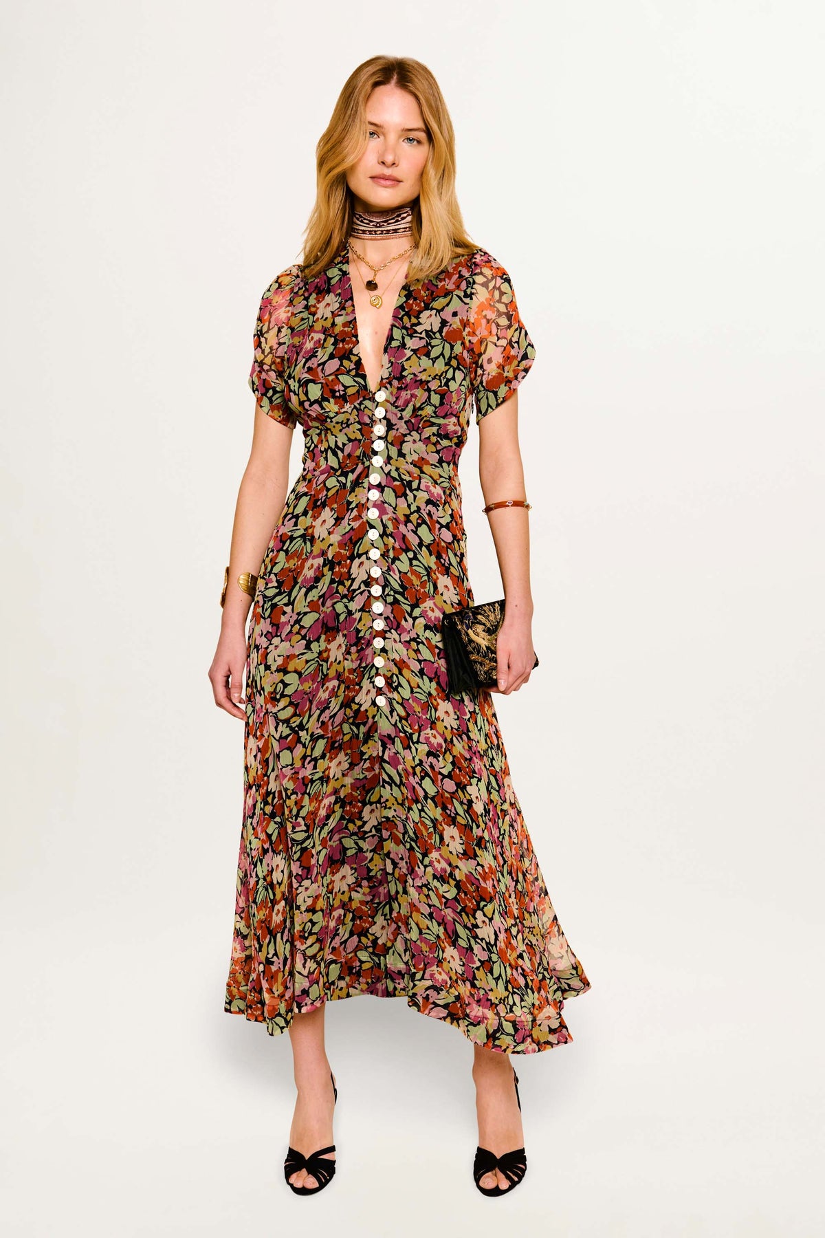 A woman wears the Novara - Vintage Floral Deep Navy dress with a deep V-neck, short sleeves, and front buttons. She completes the look with a black clutch, black heels, layered necklaces, bracelets, and a choker.