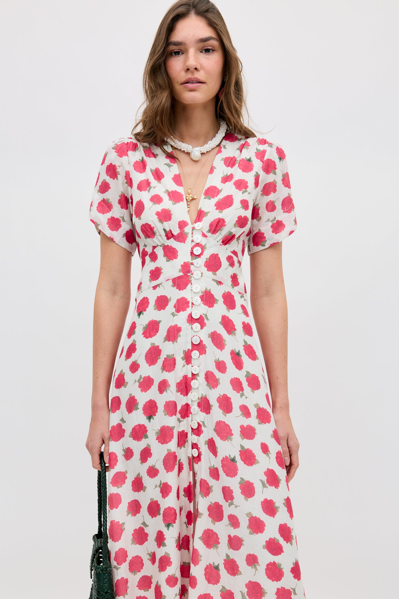 A woman stands facing forward in the Novara - Vintage Rose Ivory dress, featuring pink and green floral prints, short sleeves, and a button-down front. She accessorizes with a necklace and holds a dark bag at her side.