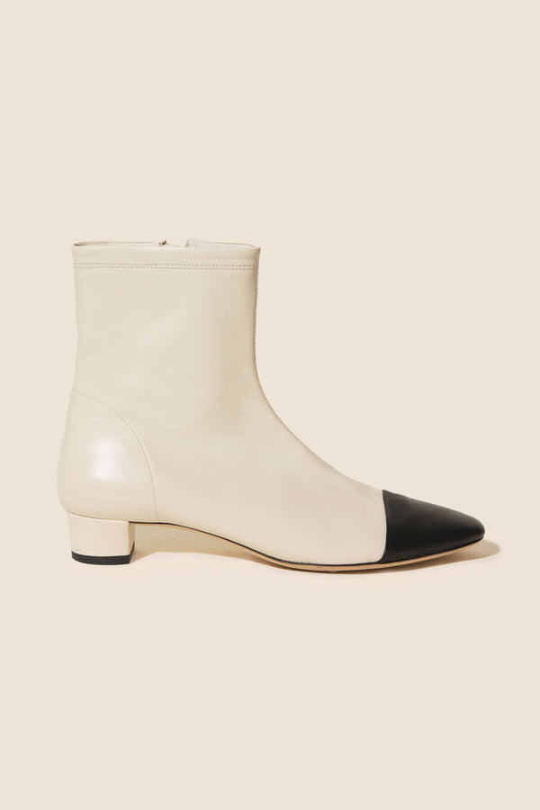 The Rey - Ivory is a dainty, feminine ankle boot in cream two-tone leather with a black pointed toe and low heel, shown in profile against a beige background.