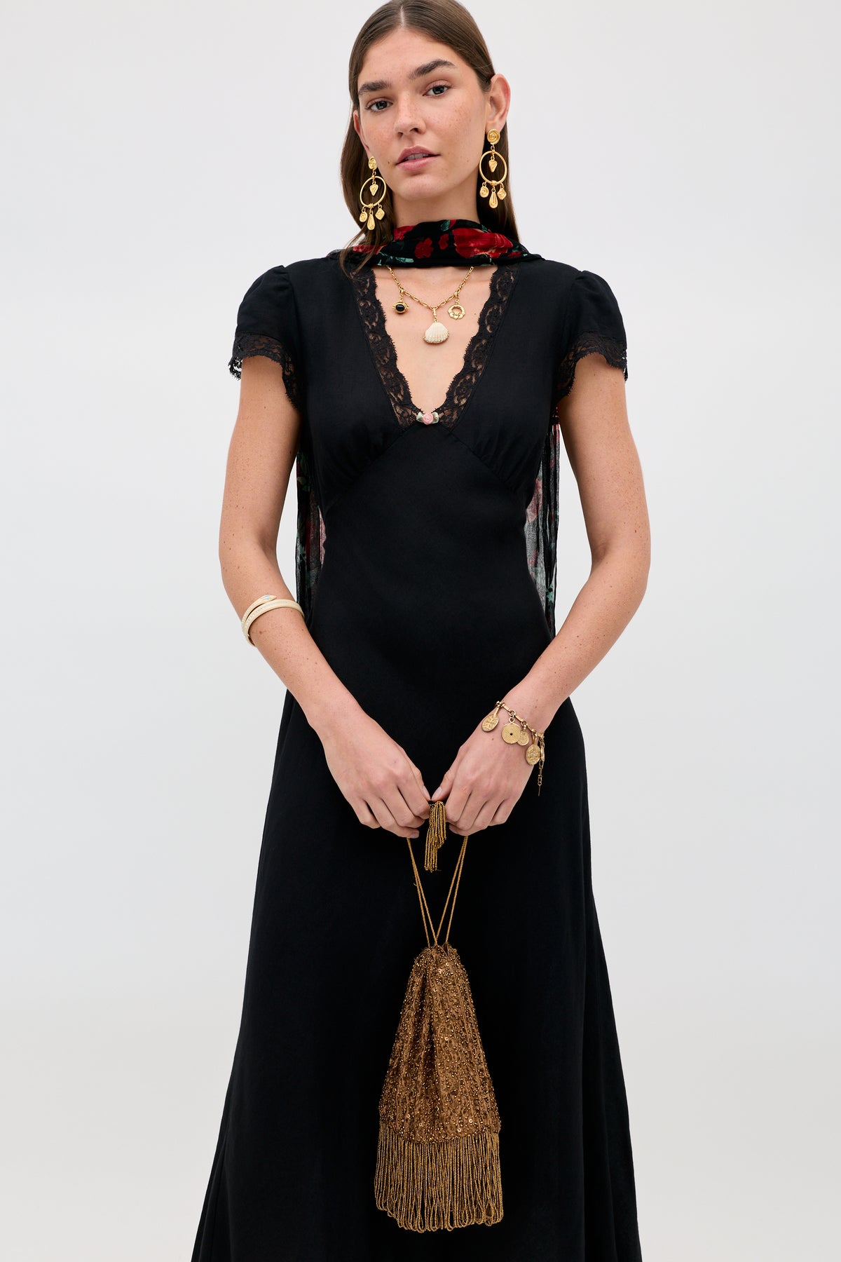 A woman wearing a black dress with lace trim, gold jewelry, a floral scarf, and holding the Orla - Vintage Scatter Embellishment Gold handbag stands against a plain light background.