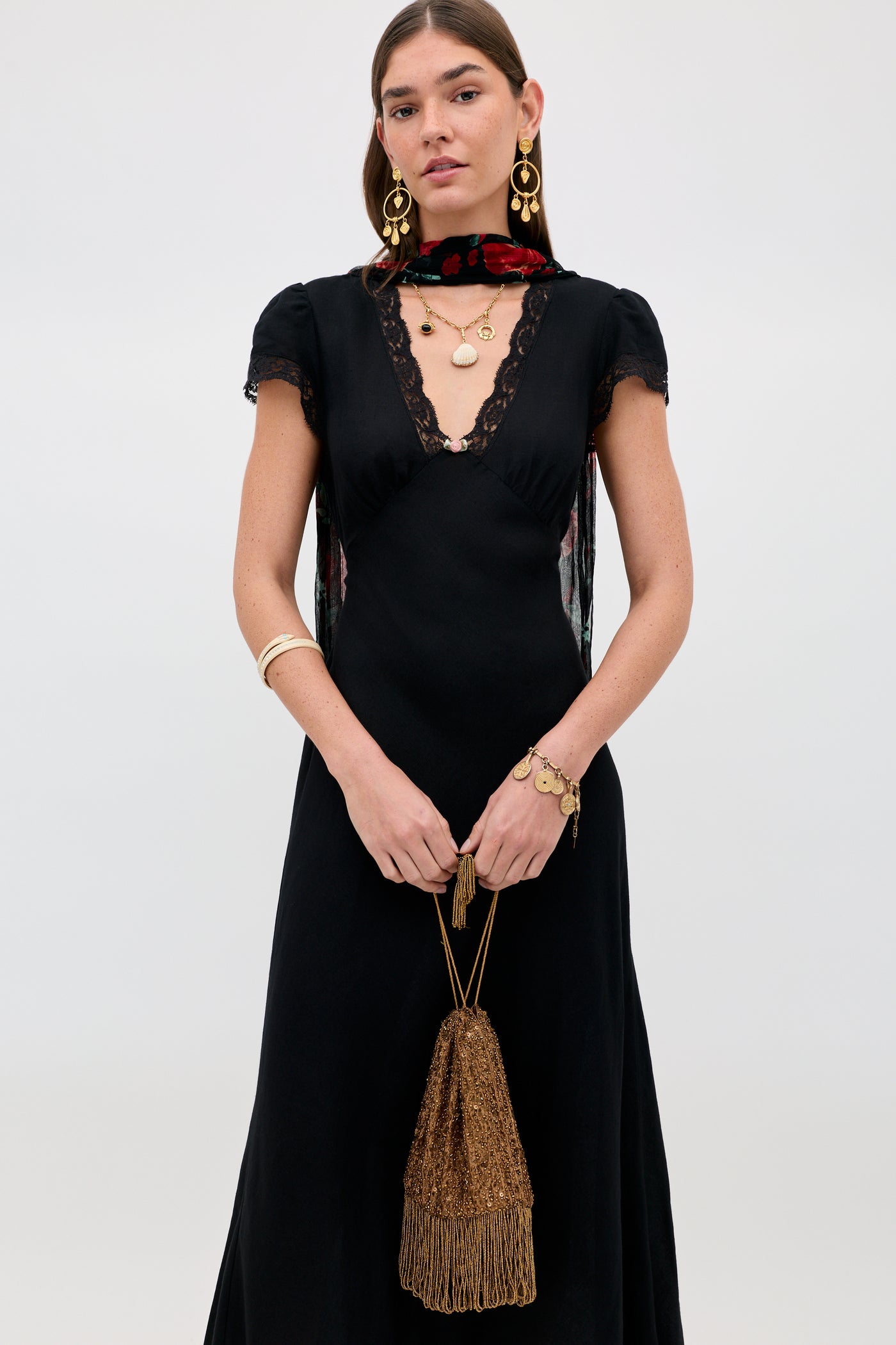 A woman wearing a black dress with lace trim, gold jewelry, a floral scarf, and holding the Orla - Vintage Scatter Embellishment Gold handbag stands against a plain light background.
