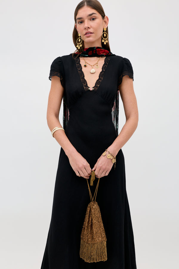 A woman wearing a black dress with lace trim, gold jewelry, a floral scarf, and holding the Orla - Vintage Scatter Embellishment Gold handbag stands against a plain light background.