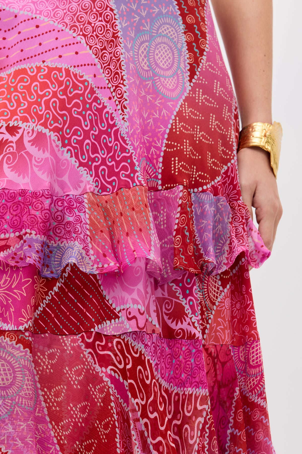 A person wears the Preslee - Ibiza Night Sunset dress, featuring bright red, pink, and purple patterns with a ruffled midi skirt, paired with a chunky gold bracelet.