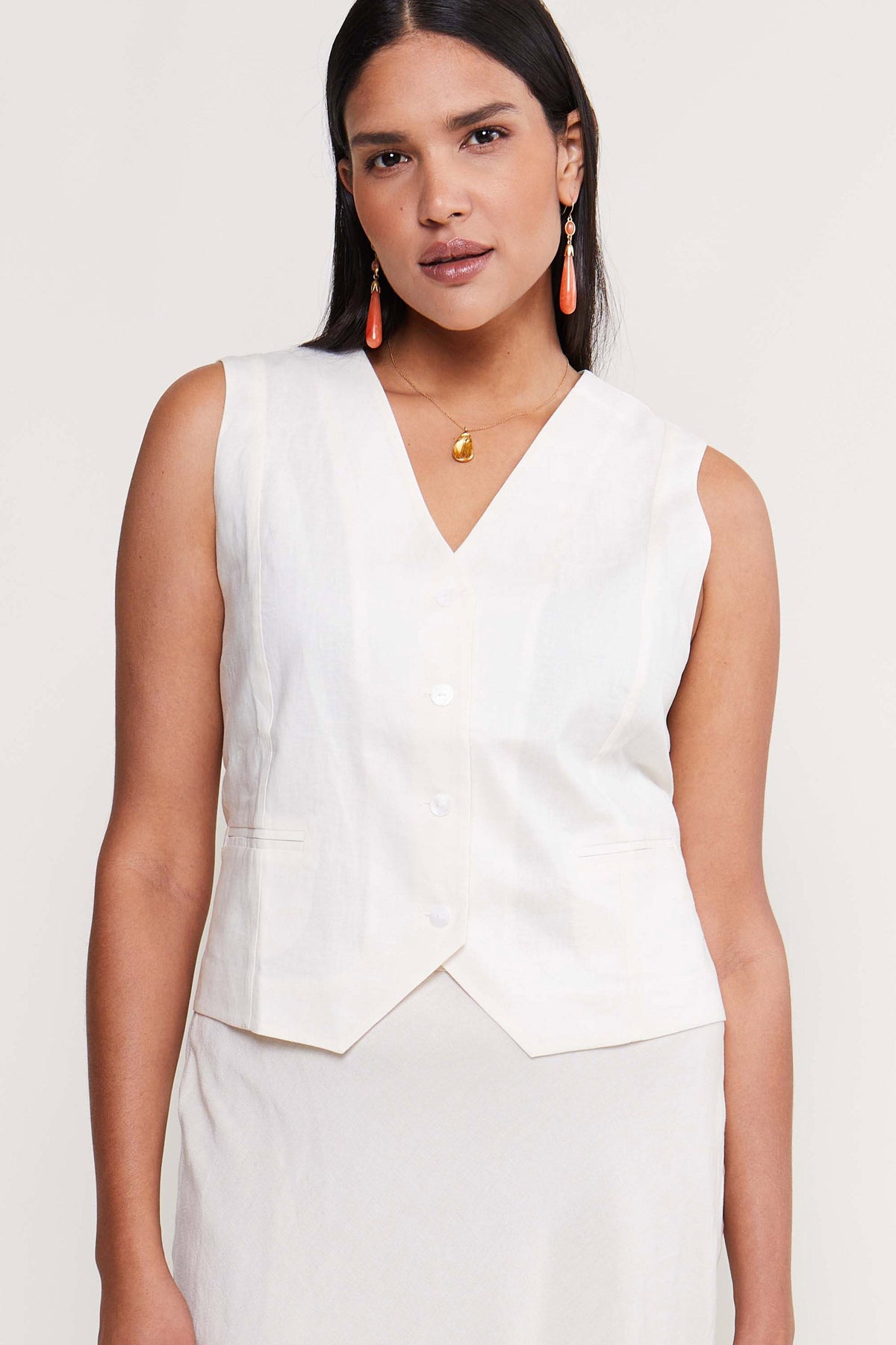 A woman with long dark hair wears the Norah - Cream sleeveless cotton-linen top and matching skirt, accessorized with drop earrings and a pendant necklace, standing against a light background.
