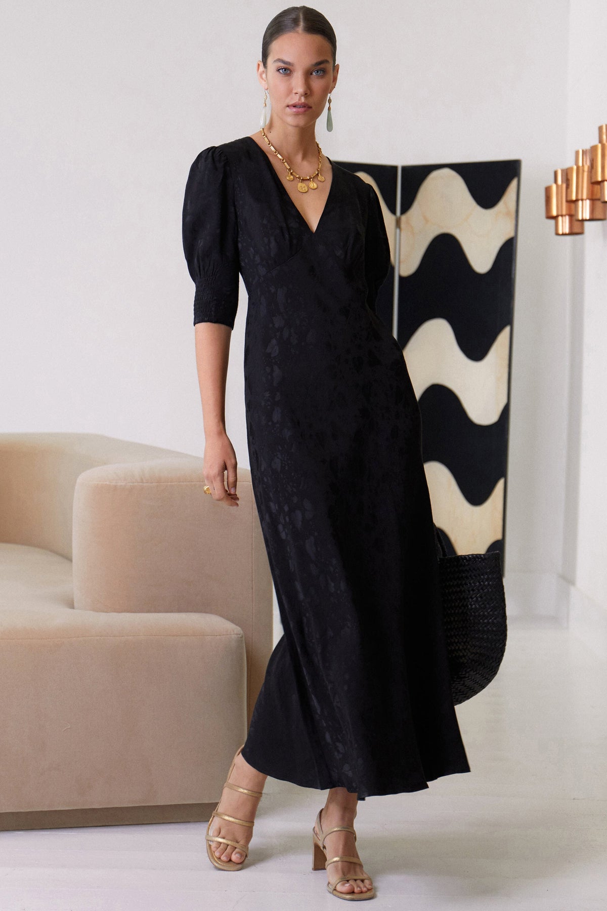 A woman stands indoors wearing the RIXO Zadie - Leaf Heart Black midi dress with puffed elbow sleeves and a V-neck. She accessorizes with gold jewelry, beige heeled sandals, and a black textured bag. Modern furniture is in the background.